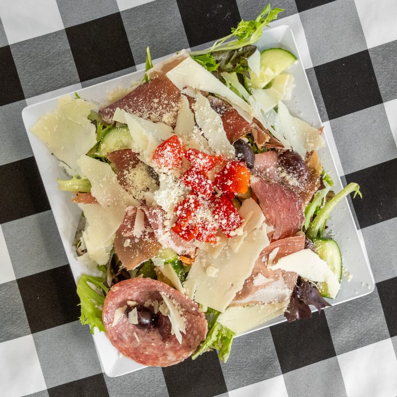 Antipasto salad with salami, prosciutto, cheese, olives, and red peppers on a checkered tablecloth.