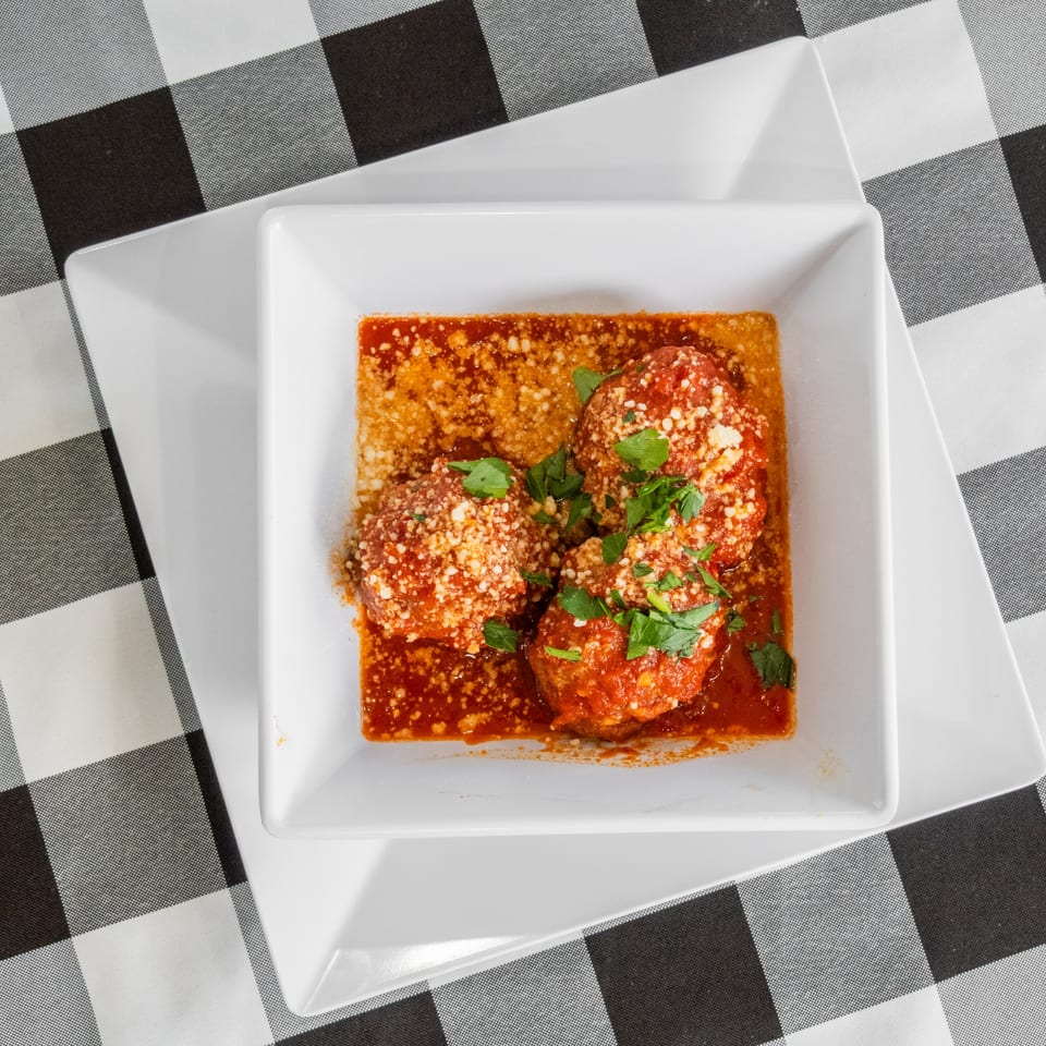 Three meatballs in rich red sauce, topped with Parmesan cheese and fresh parsley, on a white square plate.